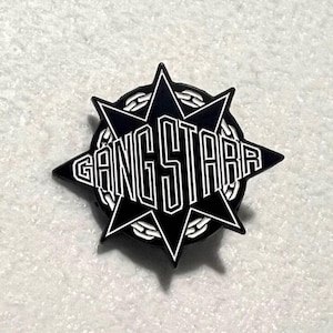 Puede incluir: Un pin de esmalte negro y blanco con el texto "GANG STARR". El diseño incluye una forma de estrella con bordes puntiagudos y un borde de cadena. El pin está sobre un fondo gris claro.
