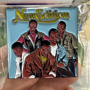 May include: A rectangular pin badge featuring a group of five men. The pin has a light blue background with the words "New Edition" in yellow script. The men are depicted in a stylized, cartoonish manner, wearing jackets and shirts in various colours.