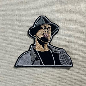 May include: Embroidered patch featuring a stylized portrait of a person wearing a fedora and a jacket. The patch has a black border, with the person's face and jacket in shades of tan, black, and gray. The hat is black and gray.