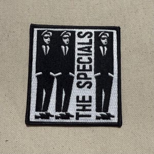 Op de afbeelding: Zwart-wit geborduurde patch met drie gestileerde figuren in pakken en hoeden. De tekst "THE SPECIALS" is verticaal uitgelijnd. De patch heeft een zwarte rand. Ideaal voor jassen, tassen of andere stoffen items.