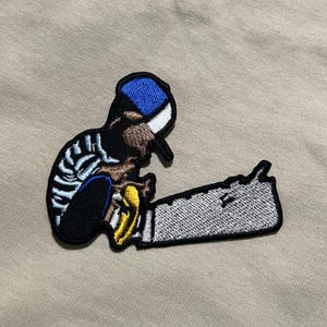 May include: Embroidered patch featuring a cartoon character with a blue and white hat, black jacket, and a chainsaw. The patch has a black border and is set against a tan background. The chainsaw has a silver blade and yellow handle.