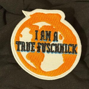 May include: A circular embroidered patch featuring an orange globe design with a white border. The text "I AM A TRUE FUSCHNICK" is stitched in black with a light blue outline across the center of the orange globe graphic.
