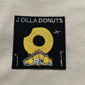 May include: Black square patch with the text "J Dilla Donuts" above a yellow donut with a bite taken out of it. Below the donut is a yellow building with a brown roof. A small airplane flies in the sky.