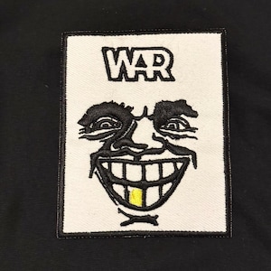 May include: A rectangular embroidered patch featuring a white background with a black border. The design displays the word WAR in black letters above a stylized black-and-white face with a wide, toothy grin and a single prominent yellow tooth.
