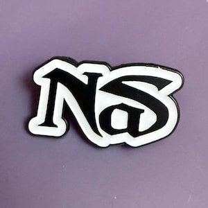 Op de afbeelding: Zwart-witte emaille pin met het woord "Nas" in een gestileerd lettertype. De pin heeft een zwarte basis met een witte omtrek, tegen een paarse achtergrond.