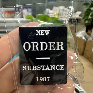 Könnte beinhalten: Ein schwarzer rechteckiger Anstecker mit den weißen Worten "NEW ORDER SUBSTANCE 1987". Der Anstecker befindet sich in einer transparenten Kunststoffverpackung. Der Hintergrund ist verschwommen und zeigt einen Blick auf eine Pflanze und ein Fenster.