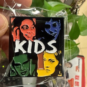 May include: A rectangular enamel pin with four coloured faces (red, blue, green, and yellow) on a black background. The word "KIDS" is in white block letters. The pin is in a clear plastic package.