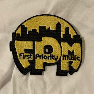 May include: Embroidered patch with a black border and a yellow interior. The patch features the letters "FPM" in yellow, with the words "First Priority Music" below. A black cityscape silhouette is at the top of the patch.