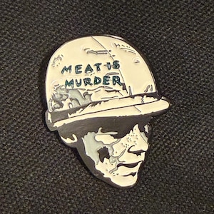 May include: A black and off-white enamel pin featuring a graphic illustration of a person wearing a helmet. The text MEAT IS MURDER is clearly printed in dark lettering across the front of the helmet, with a high-contrast design throughout.