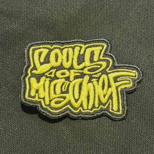 Può includere: Una toppa ricamata gialla con le parole "SOULS OF MISCHIEF" in un font stilizzato. La toppa ha un bordo nero ed è posta su uno sfondo di tessuto verde scuro.