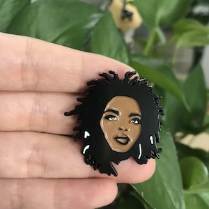 May include: A black enamel pin depicting a woman with dark hair and brown skin. The pin has a detailed design with white accents. It is likely a collectible item or accessory.