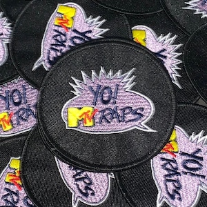 May include: Black round embroidered patches with the text "YO! MTV RAPS" in a speech bubble. The MTV logo is yellow and red. The text and speech bubble are purple with white accents. Patches are a retro style.