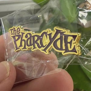May include: A gold-colored pin with the text "The Pharcyde" in purple lettering. The pin is in a clear plastic bag, held by a person. The pin has a shiny, metallic finish and a decorative font.