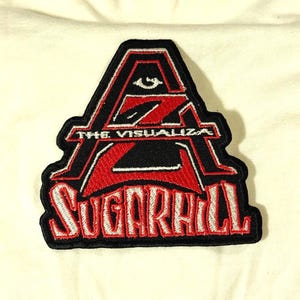 Puede incluir: Un parche bordado en negro, rojo y blanco que presenta una letra A estilizada con un símbolo de ojo en la parte superior. El texto THE VISUALIZA está centrado dentro de la letra, y la palabra SUGARHILL está bordada en letras blancas grandes.