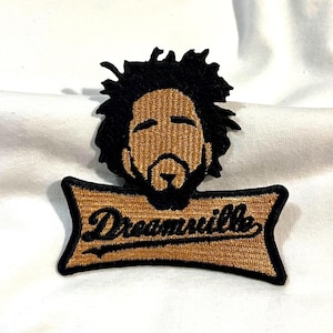 May include: Embroidered patch featuring a stylized portrait with black hair and beard. Below the portrait is a banner with the word "Dreamville" in a script font. The patch has a gold and black color scheme.