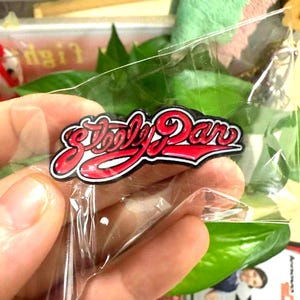 Könnte beinhalten: Ein roter und weißer Steely Dan-Anstecker mit schwarzem Rand wird in einer transparenten Plastikverpackung gehalten. Der Anstecker zeigt das Logo der Band in einer stilisierten Schrift. Der Hintergrund umfasst grüne Blätter und andere Gegenstände.