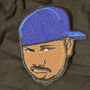 May include: An embroidered patch featuring a stylized portrait of a person with a brown complexion, a dark beard, and a mustache, wearing a bright blue baseball cap. The patch has a black border and is set against a solid black fabric background.