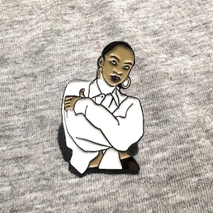May include: Enamel pin featuring a stylized illustration of a person in a white shirt and gold hoop earrings. The pin has a black outline and is set against a gray background.