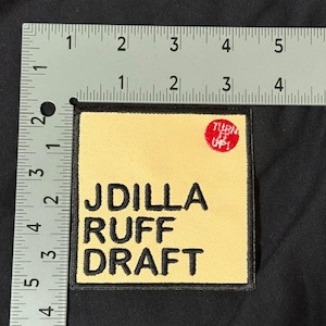 May include: A square embroidered patch with a light yellow background and a black border. The text reads JDILLA RUFF DRAFT in black, with a small red circle containing the white text TURN IT UP! A metal ruler shows the patch is 7.6 cm wide.