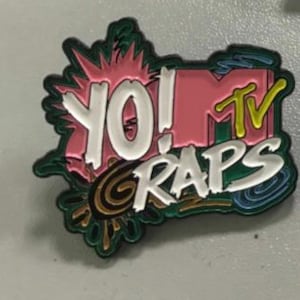 May include: A colorful enamel pin featuring the text "YO! MTV RAPS" in white, pink, and yellow. The pin has a retro design with green, blue, and gold accents, reminiscent of 1990s pop culture.