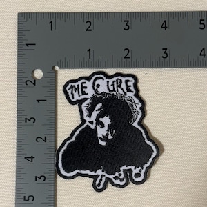 Könnte beinhalten: Ein schwarz-weiß bestickter Aufnäher mit dem Porträt einer Person mit zerzaustem Haar und dem Text THE CURE darüber. Der Aufnäher liegt neben einem Metalllineal und ist etwa 7,6 cm hoch und 6,4 cm breit.