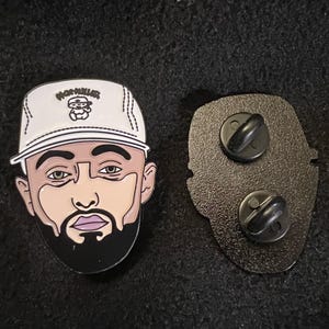 May include: A black and white enamel pin featuring a stylized illustration of a person wearing a white baseball cap with the text "MORNILEZ". The pin has a black beard and a pink mouth. The back of the pin has two black pin backs.