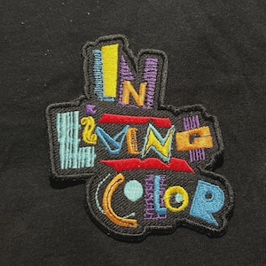 以下が含まれることがあります： カラフルなレトロデザインで「IN LIVING COLOR」の文字が刺繍されたパッチ。パッチは黒い縁取りがあり、黒い背景に配置されています。テキストは、黄色、紫、赤、青など、さまざまな色で表示されています。