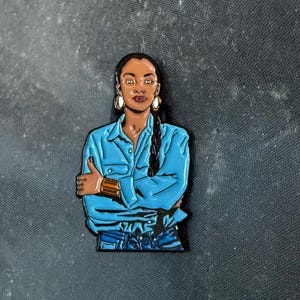 May include: Enamel pin featuring a stylized illustration of a person with dark skin, wearing a blue button-down shirt and jeans. The pin has gold-colored hoop earrings and a cuff bracelet. The person has their arms crossed.