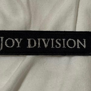 May include: A black rectangular patch with the embroidered text "JOY DIVISION" in silver. The patch is likely for clothing or accessories, and is set against a white background.