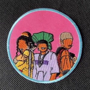 May include: Embroidered patch featuring three stylized figures in a circle. The patch has a pink background with a light blue border. The figures are in yellow, orange, and purple clothing. The patch is likely for clothing or accessories.