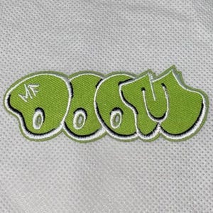Può includere: Una toppa ricamata verde lime con la scritta "DOOM" in un font graffiti stilizzato. La toppa ha un bordo bianco e le lettere hanno un effetto 3D. Le lettere "MF" sono nella prima lettera.