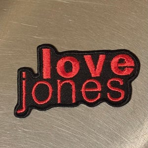 Può includere: Toppa nera ricamata con le parole "love jones" in rosso. La toppa ha un bordo nero e una forma rettangolare con angoli arrotondati. Il testo è in grassetto, carattere sans-serif.
