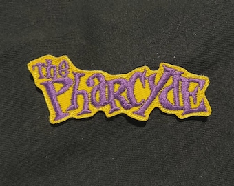 May include: A yellow embroidered patch featuring the purple text "The Pharcyde", with a yellow border. The patch is set against a black background. The text is in a decorative font.
