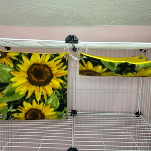 Puede incluir: Un accesorio cuadrado para jaulas de mascotas y una hamaca a juego, ambos con un dise&ntilde;o de girasol. El accesorio cuadrado muestra girasoles amarillos sobre un fondo negro. La hamaca es amarilla con un estampado de girasoles.