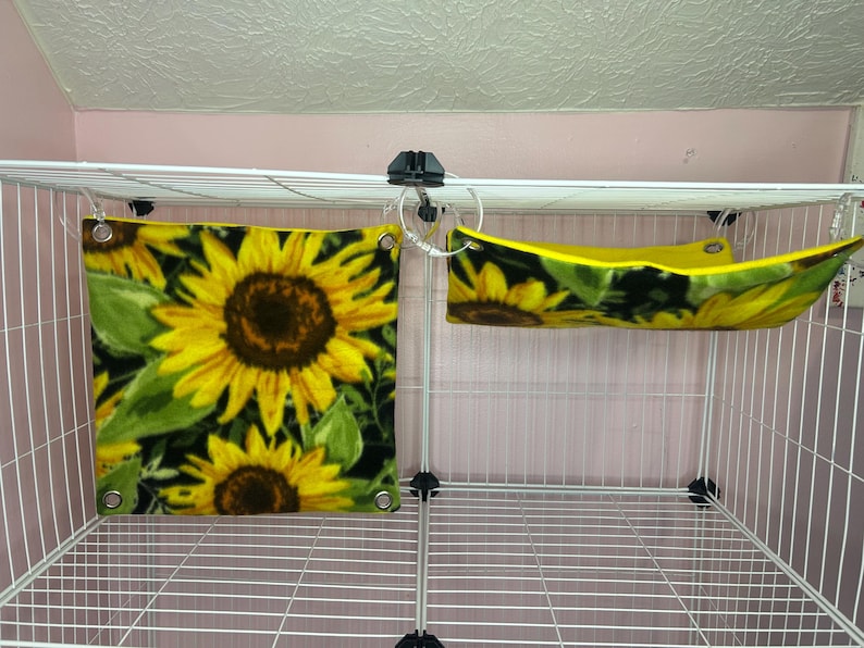 Puede incluir: Un accesorio cuadrado para jaulas de mascotas y una hamaca a juego, ambos con un dise&ntilde;o de girasol. El accesorio cuadrado muestra girasoles amarillos sobre un fondo negro. La hamaca es amarilla con un estampado de girasoles.