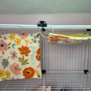 Puede incluir: Una jaula de alambre blanca con dos accesorios de forro polar. Uno es un coj&iacute;n cuadrado con un estampado floral en naranja, rosa, amarillo y gris. El otro es una hamaca con un dise&ntilde;o floral similar.