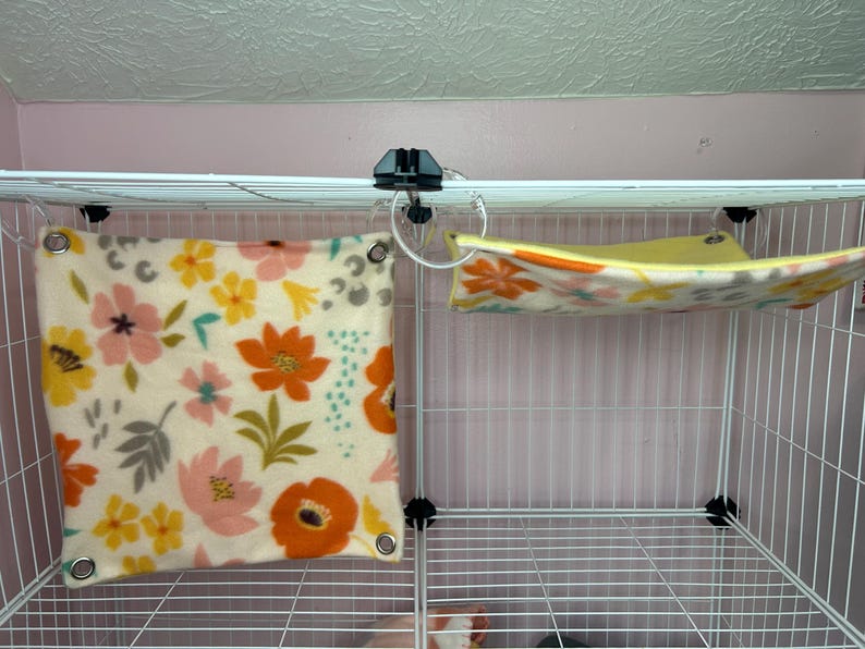 Puede incluir: Una jaula de alambre blanca con dos accesorios de forro polar. Uno es un coj&iacute;n cuadrado con un estampado floral en naranja, rosa, amarillo y gris. El otro es una hamaca con un dise&ntilde;o floral similar.