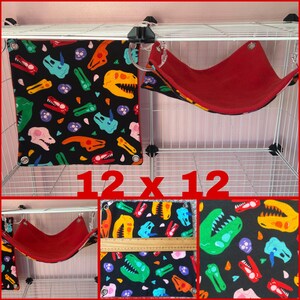 Puede incluir: Un accesorio para mascotas cuadrado negro con un colorido estampado de dinosaurios, de 30 x 30 cm, cuelga junto a una hamaca roja. El dise&ntilde;o de dinosaurios incluye varias cabezas y huesos de dinosaurios de colores. La hamaca tambi&eacute;n est&aacute; sujeta a una jaula de alambre.