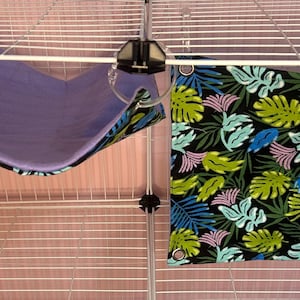 Puede incluir: Dos accesorios para mascotas: una hamaca morada y una tela negra con un estampado de hojas tropicales. La hamaca est&aacute; suspendida de una jaula de alambre. La tela tiene hojas verdes, azules y rosas.