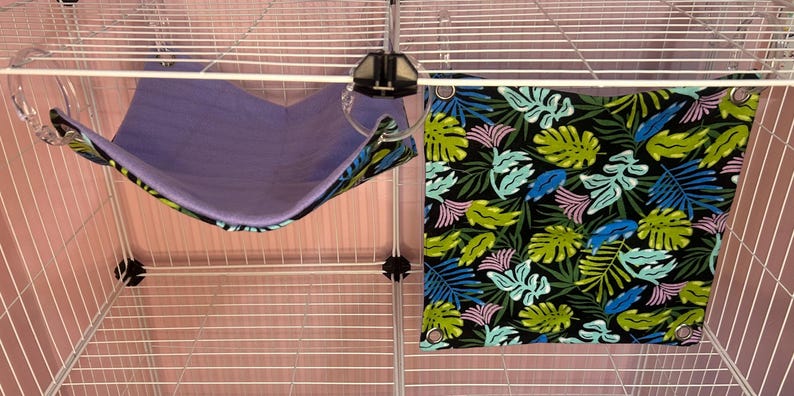 Puede incluir: Dos accesorios para mascotas: una hamaca morada y una tela negra con un estampado de hojas tropicales. La hamaca est&aacute; suspendida de una jaula de alambre. La tela tiene hojas verdes, azules y rosas.