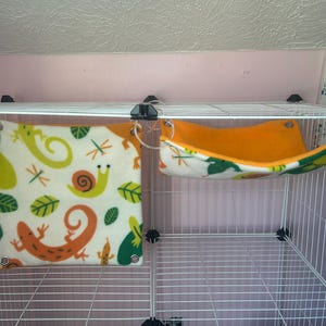 Puede incluir: Un peque&ntilde;o h&aacute;bitat para mascotas con dos accesorios de forro polar. Uno es una hamaca cuadrada de color crema con dise&ntilde;os de lagartos, caracoles y hojas de color naranja, verde y amarillo. El otro es una hamaca naranja y blanca con un estampado de hojas.