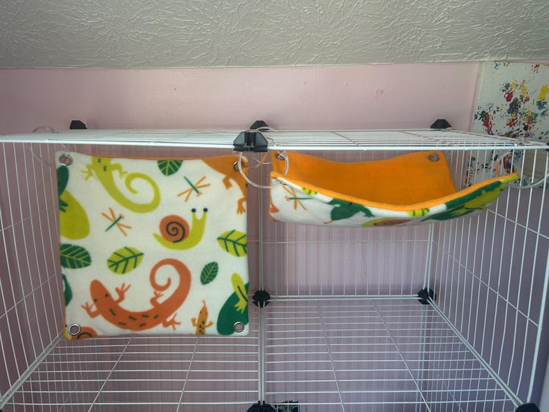 Puede incluir: Un peque&ntilde;o h&aacute;bitat para mascotas con dos accesorios de forro polar. Uno es una hamaca cuadrada de color crema con dise&ntilde;os de lagartos, caracoles y hojas de color naranja, verde y amarillo. El otro es una hamaca naranja y blanca con un estampado de hojas.