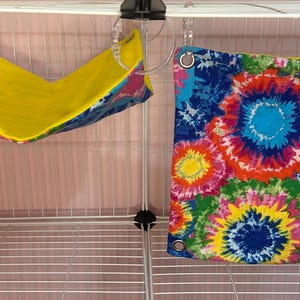 Puede incluir: Dos accesorios para mascotas: una hamaca amarilla y azul y una tela con estampado tie-dye. La hamaca est&aacute; suspendida de la parte superior de una jaula de alambre blanca. La tela tie-dye presenta una vibrante mezcla de colores, incluyendo azul, rosa, amarillo y verde.