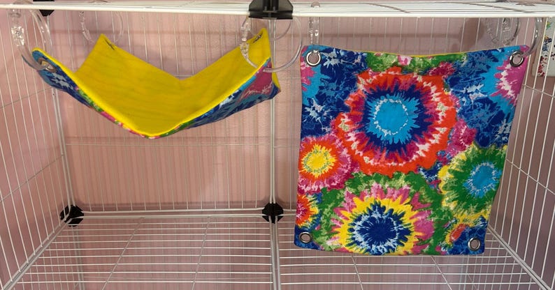 Puede incluir: Dos accesorios para mascotas: una hamaca amarilla y azul y una tela con estampado tie-dye. La hamaca est&aacute; suspendida de la parte superior de una jaula de alambre blanca. La tela tie-dye presenta una vibrante mezcla de colores, incluyendo azul, rosa, amarillo y verde.