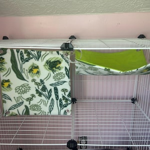 Puede incluir: Una jaula de alambre blanca con dos accesorios para mascotas. Uno es una manta cuadrada de forro polar color crema con un estampado de ranas y plantas verdes. El otro es una hamaca verde con un estampado floral.