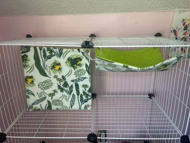 Puede incluir: Una jaula de alambre blanca con dos accesorios para mascotas. Uno es una manta cuadrada de forro polar color crema con un estampado de ranas y plantas verdes. El otro es una hamaca verde con un estampado floral.