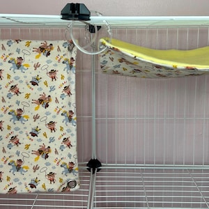 Puede incluir: Una jaula de alambre blanca con dos accesorios para mascotas. Un coj&iacute;n cuadrado de tela color crema con estampado de vaquero cuelga a la izquierda. Una hamaca amarilla con estampado floral est&aacute; suspendida a la derecha.