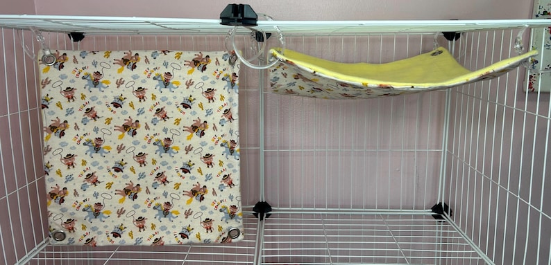 Puede incluir: Una jaula de alambre blanca con dos accesorios para mascotas. Un coj&iacute;n cuadrado de tela color crema con estampado de vaquero cuelga a la izquierda. Una hamaca amarilla con estampado floral est&aacute; suspendida a la derecha.
