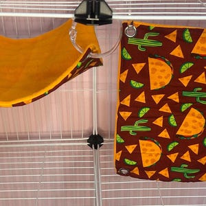Puede incluir: Una peque&ntilde;a jaula para mascotas con dos accesorios colgantes. Uno es una hamaca amarilla con un ribete estampado. El otro es una tela marr&oacute;n con un estampado de tacos, gajos de lima y cactus. Ambos est&aacute;n dise&ntilde;ados para mascotas peque&ntilde;as.