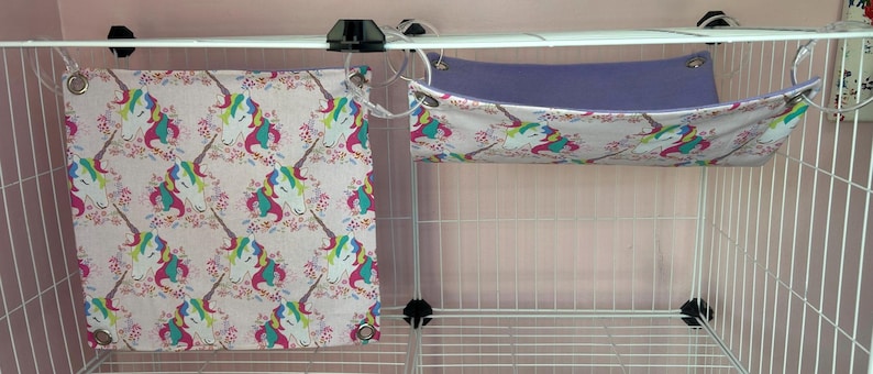 Puede incluir: Dos accesorios para mascotas: un panel de tela rectangular y una hamaca. Ambos est&aacute;n decorados con un patr&oacute;n repetido de unicornios con crines coloridas y detalles florales sobre un fondo rosa claro. La hamaca tiene un interior morado.